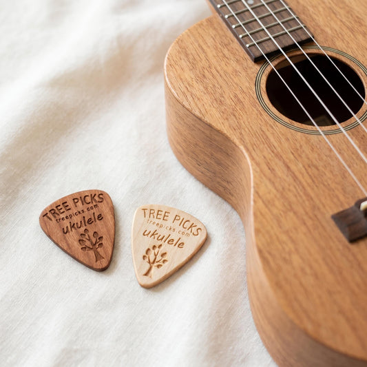 Two wooden ukulele picks with 'Tree Picks' branding next to a ukulele on a white background.